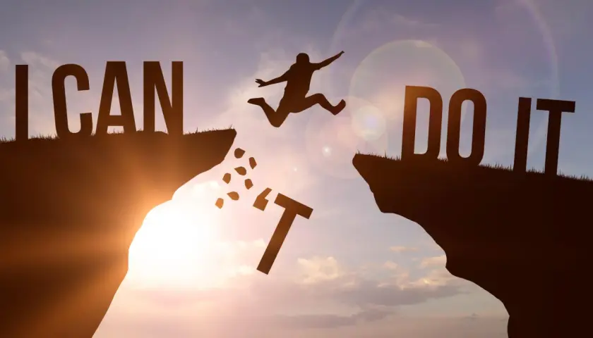 Sylwetka skaczącego człowieka nad przepaścią, litery "I CAN DO IT" na tle zachodzącego słońca.